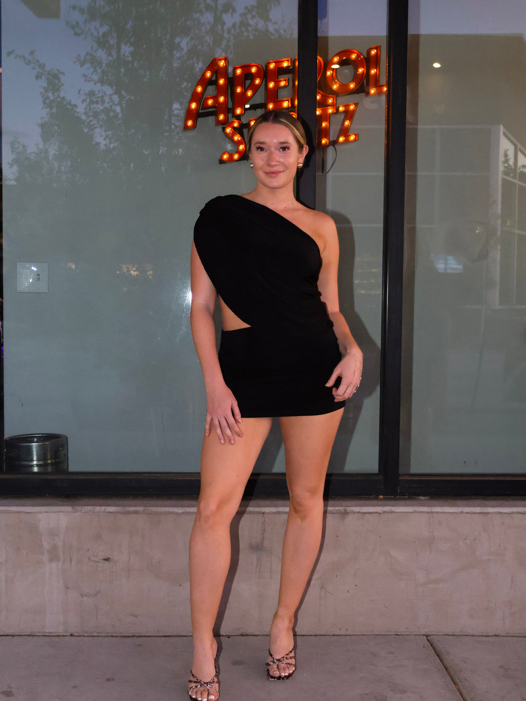 Woman in a black one-shoulder dress standing in front of a building with a neon sign.