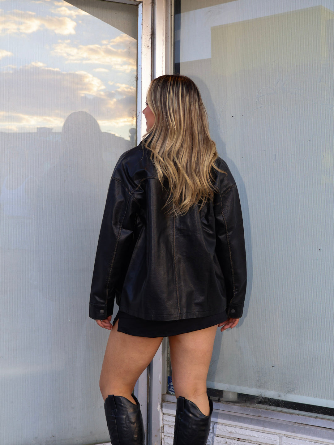Person wearing a black leather jacket and boots standing by a glass door with a reflective surface.