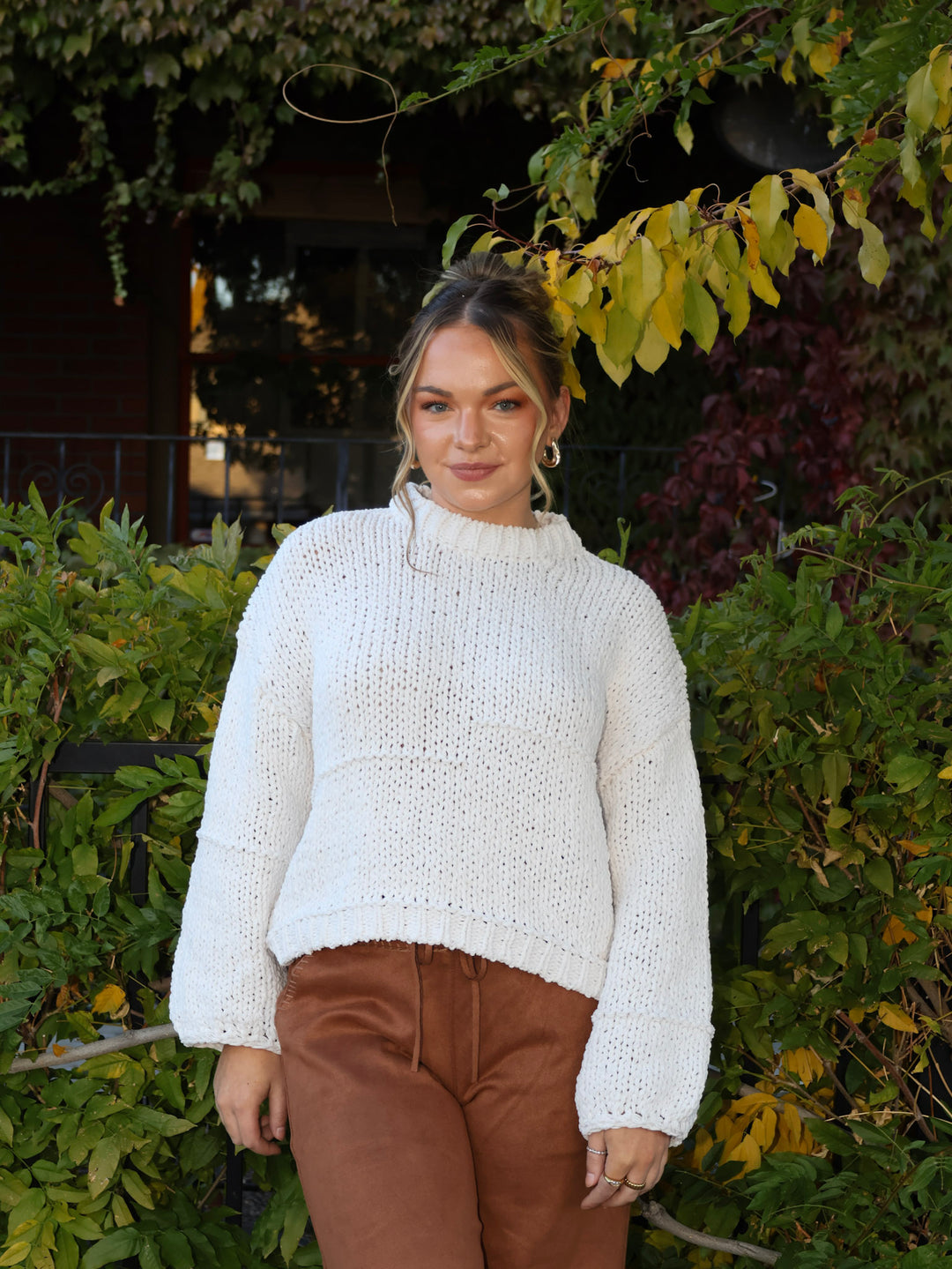 Woman wearing a white sweater and brown pants standing in front of green foliage