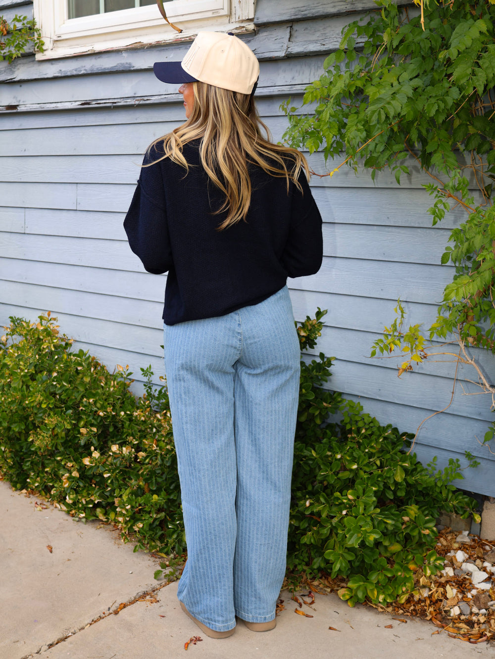 Person wearing a black jacket, light blue jeans, and a beige cap standing against a light blue wall with greenery.