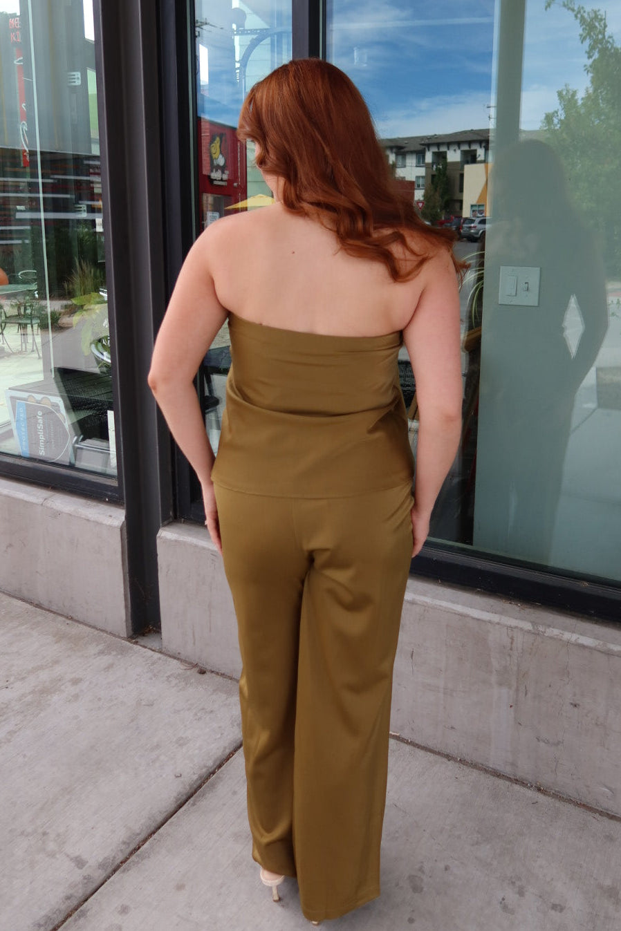 A woman with red hair is shown from behind wearing a strapless olive-brown jumpsuit while standing on a sidewalk in front of a storefront window.