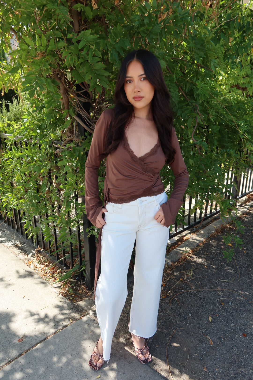 A woman stands outdoors on a sidewalk in front of green bushes and a black metal fence, wearing a brown long-sleeve wrap-style top, white wide-leg cropped jeans, and brown patterned flat sandals.