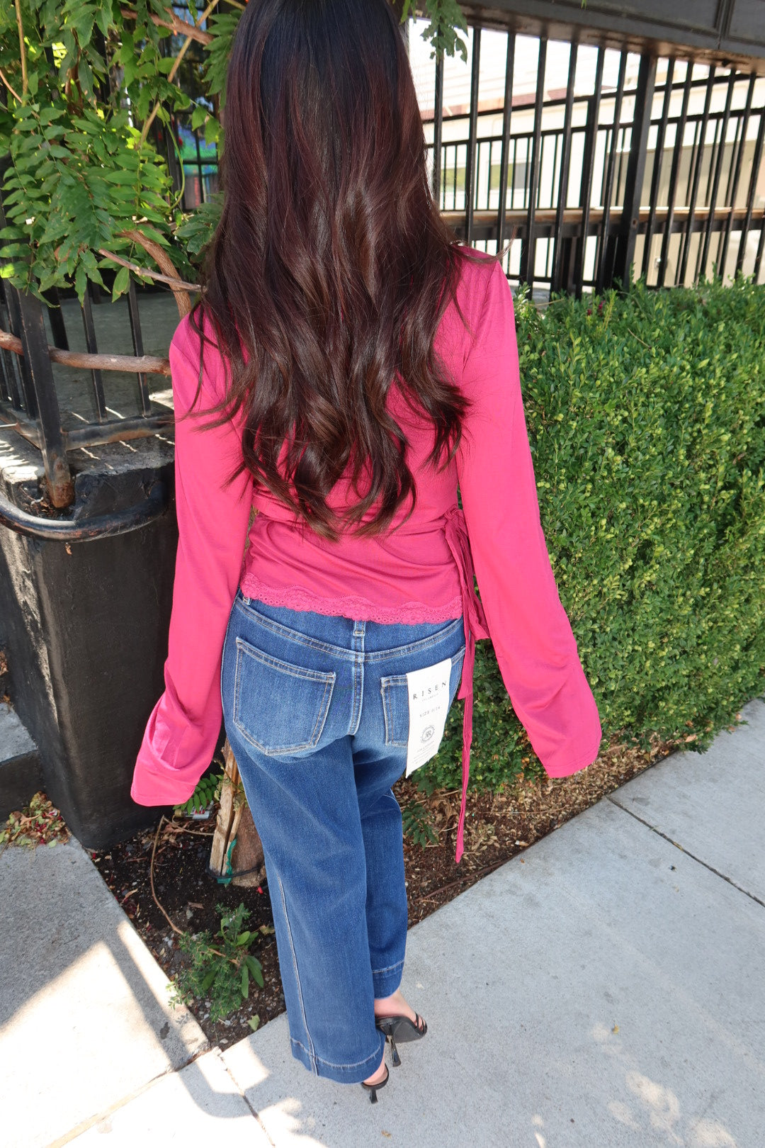 A woman viewed from behind wearing a bright pink long-sleeve top and blue jeans, standing outdoors on a sidewalk next to a green hedge.