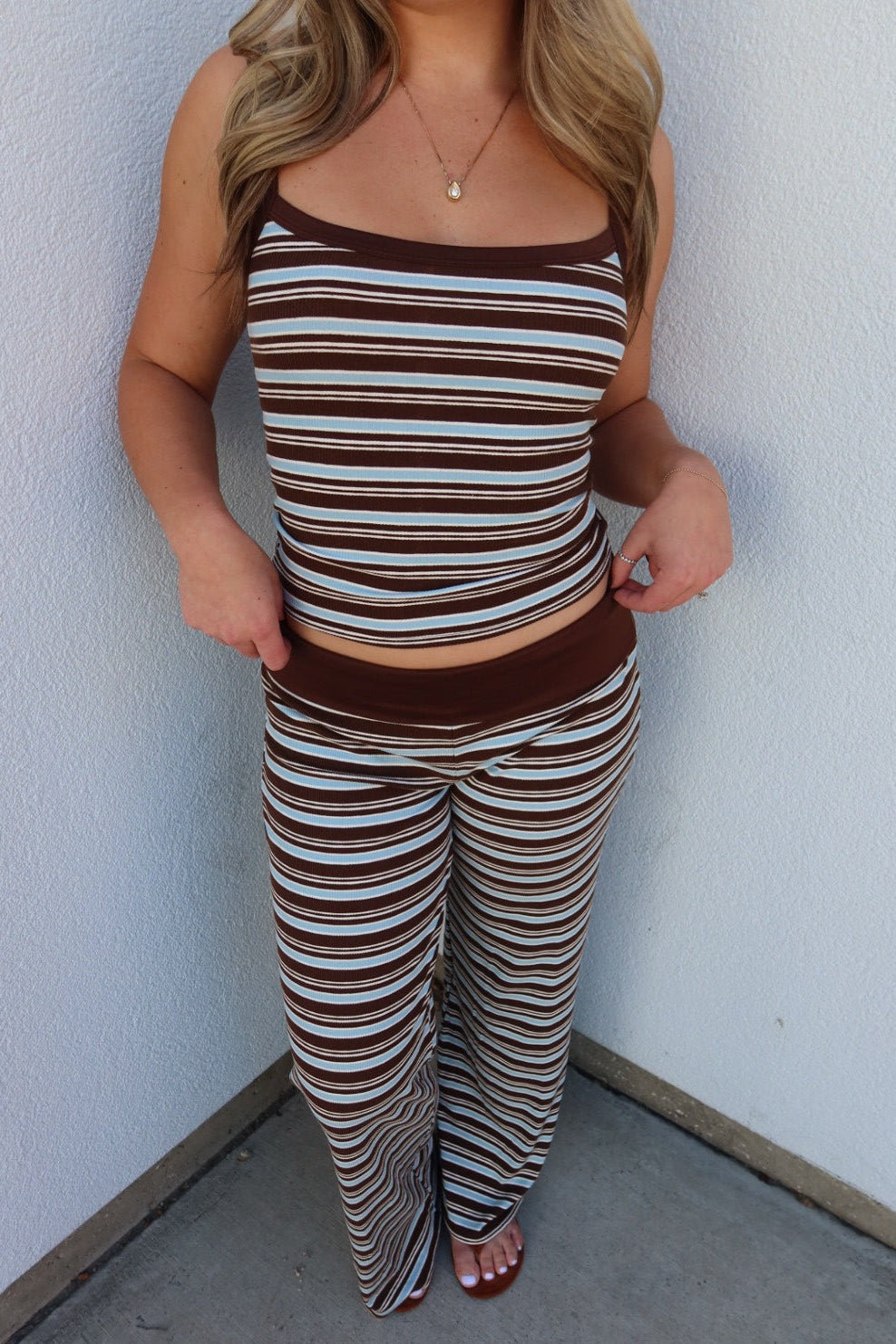 A woman wearing a matching two-piece lounge set with brown, beige, and white horizontal stripes, standing in a corner against a white wall.