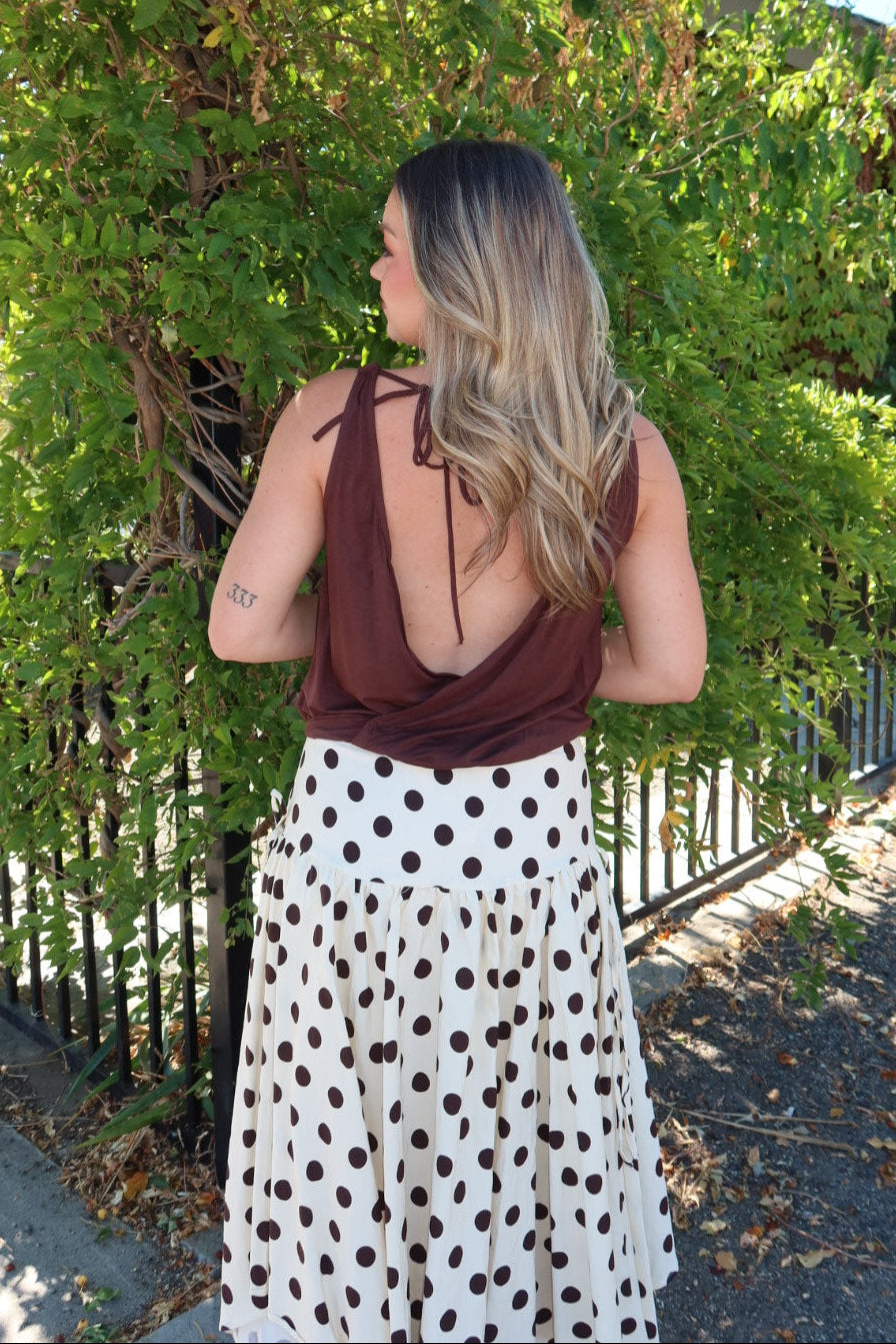 A woman stands outdoors in front of dense green foliage, wearing a brown halter-style top with a low open back and a white midi skirt with dark brown polka dots.
