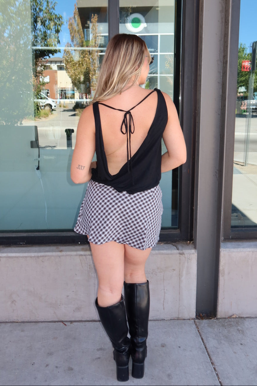A woman stands with her back to the camera in front of a storefront window, wearing a black sleeveless top with a tie-back design, a black and white gingham mini skirt, and black knee-high boots.