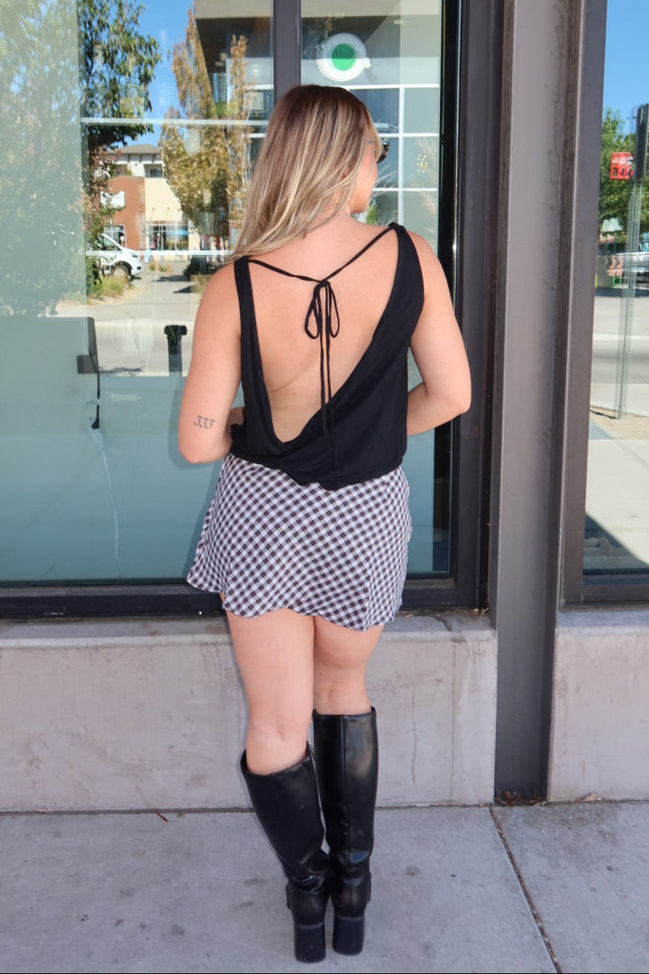 A woman stands with her back to the camera in front of a storefront window, wearing a black sleeveless top with a tie-back design, a black and white gingham mini skirt, and black knee-high boots.