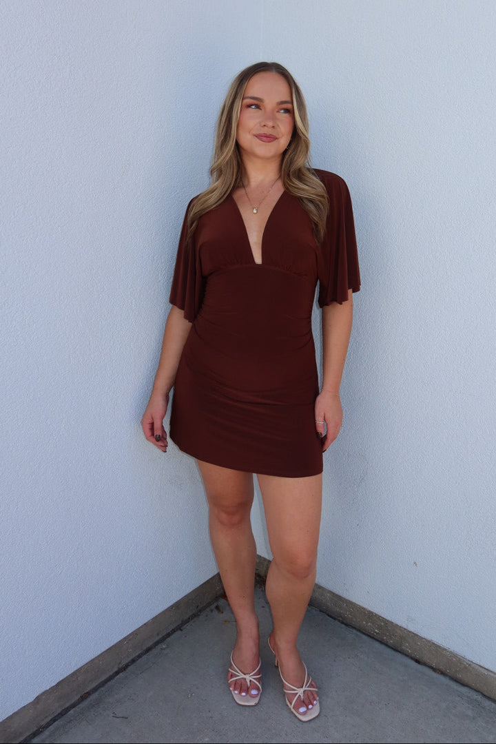 A woman modeling a short, deep brown mini dress with short flutter sleeves and a split V-neckline, standing against a plain light blue-gray wall.