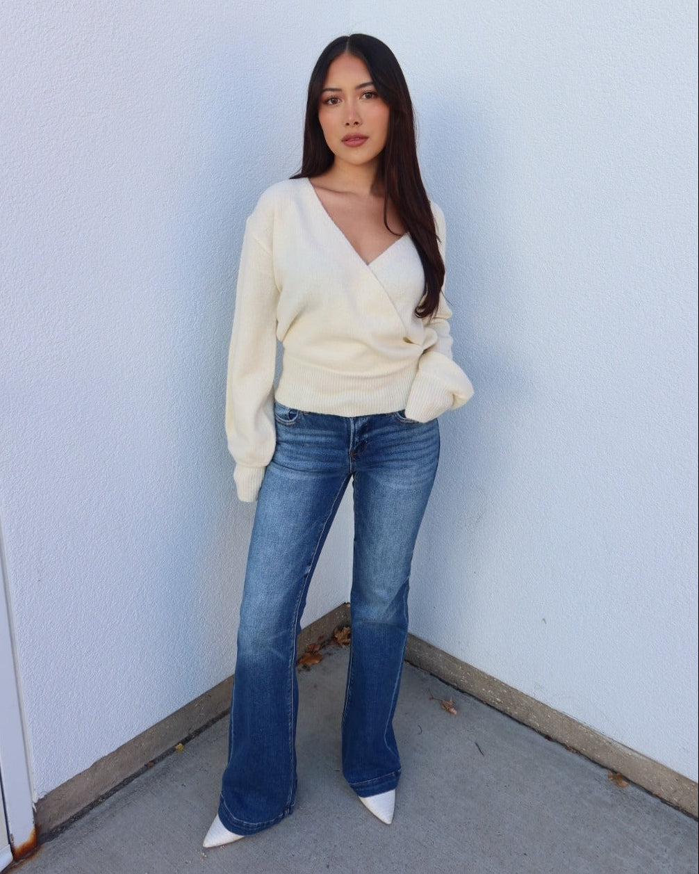 A woman stands against a light blue wall wearing a cream-colored wrap-style knit sweater, blue flare jeans, and white pointed-toe heels.