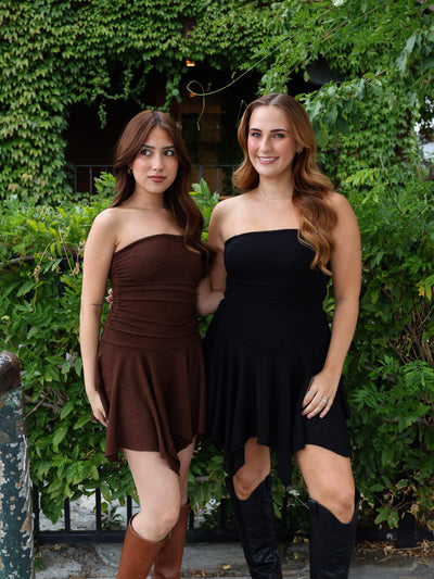 Two women wearing strapless dresses standing in front of green foliage.