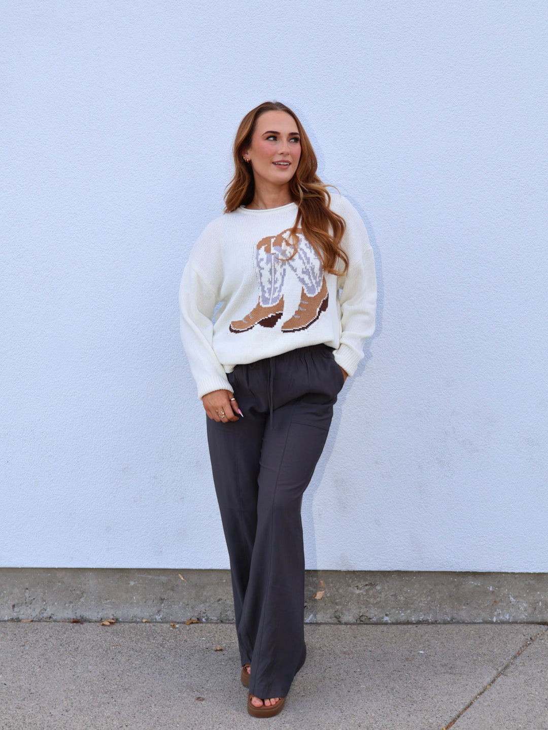 Woman wearing a white sweater with a graphic design and dark pants against a light blue wall.