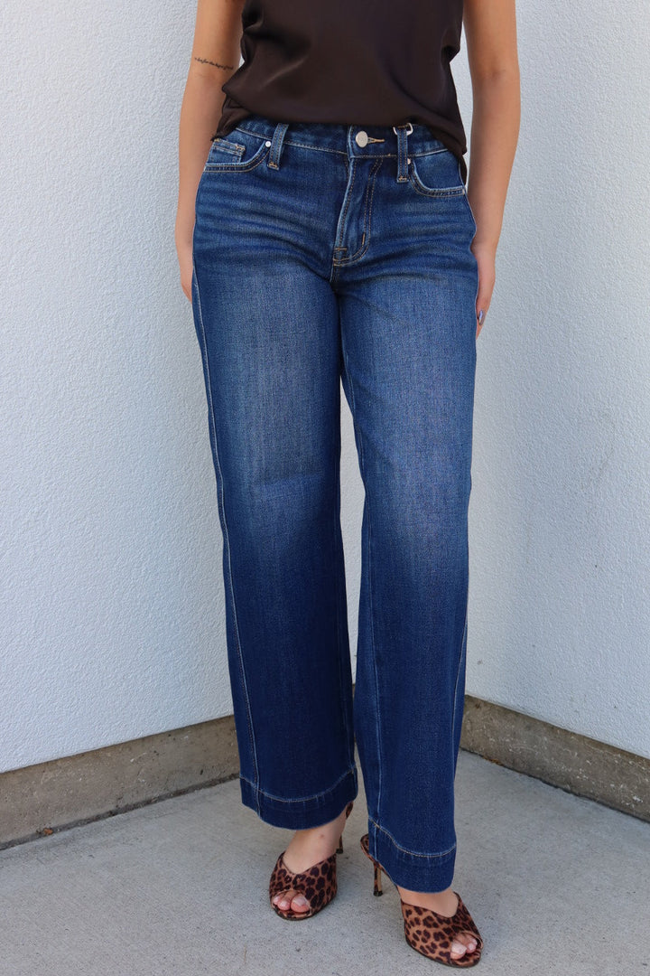 A person wearing dark blue wide-leg jeans, a dark brown sleeveless top, and leopard-print heeled sandals, standing against a light gray textured wall.