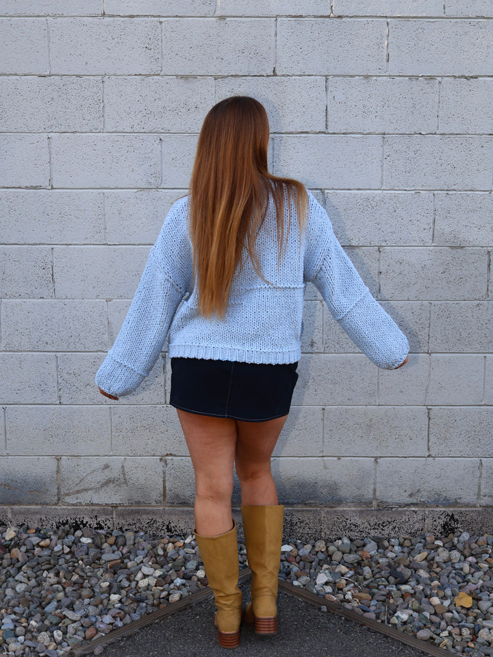 Person wearing a light blue sweater and black skirt standing against a gray brick wall.