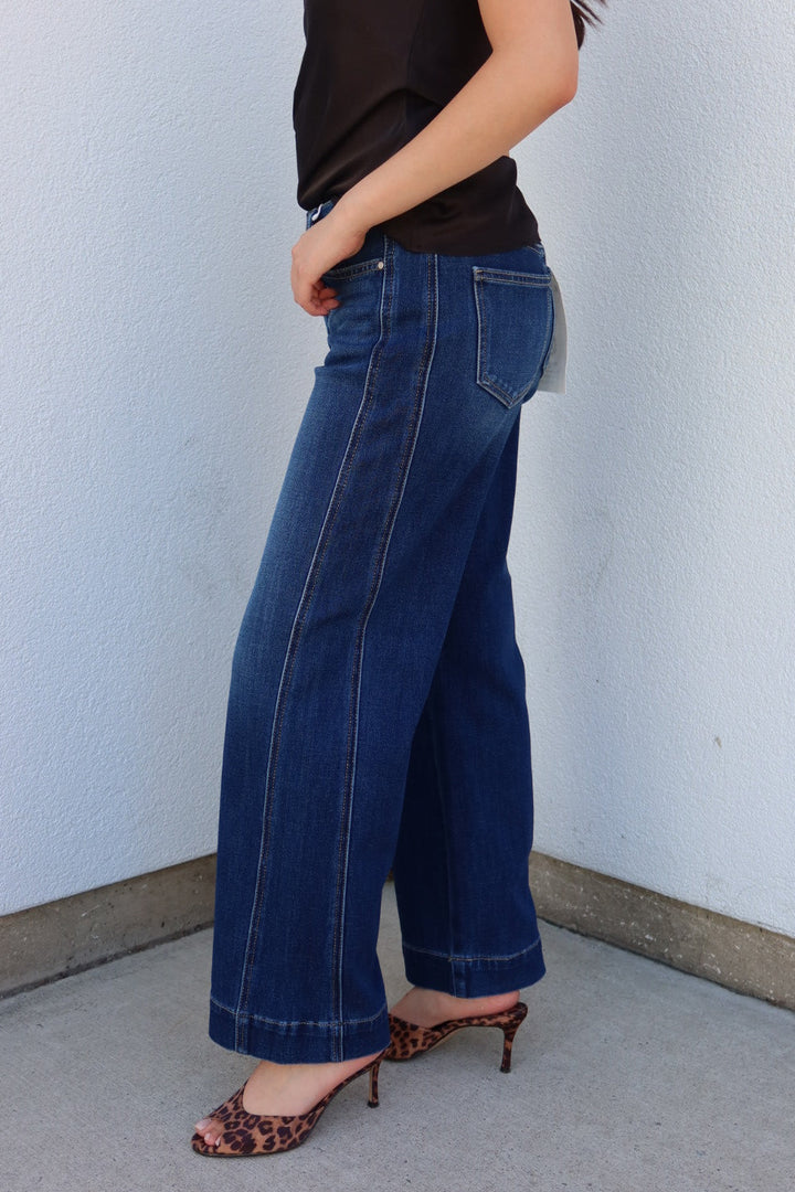 A person wearing dark blue wide-leg cropped jeans, a black sleeveless top, and leopard-print heeled mules, standing against a light gray wall.