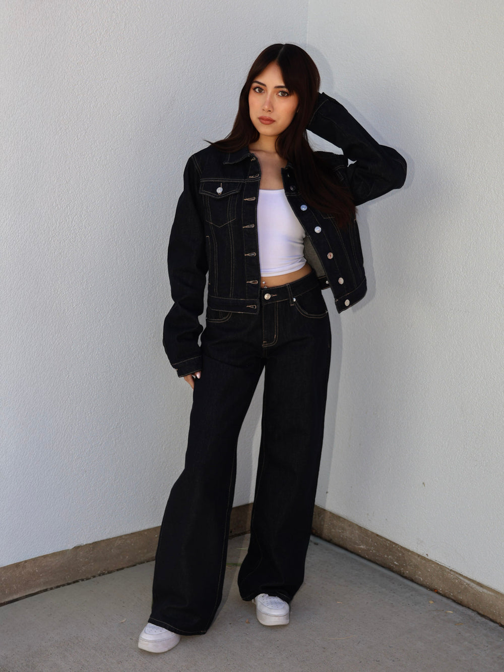 Person wearing a black denim jacket and black flared jeans against a plain wall.