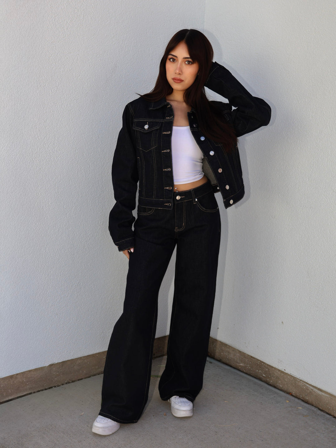 Person wearing a black denim jacket and black flared jeans against a plain wall.