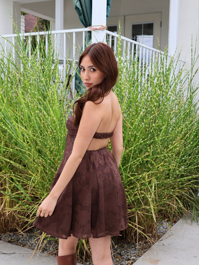 Woman in a brown dress standing outdoors with greenery in the background