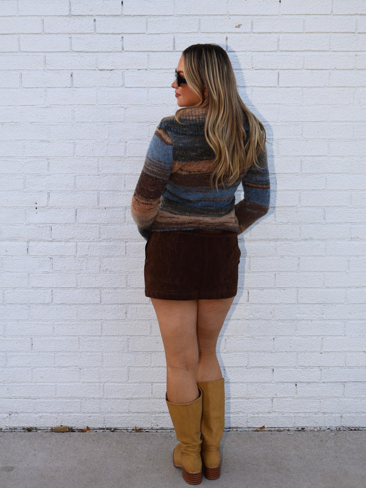 Woman wearing a striped sweater and brown skirt against a white brick wall.