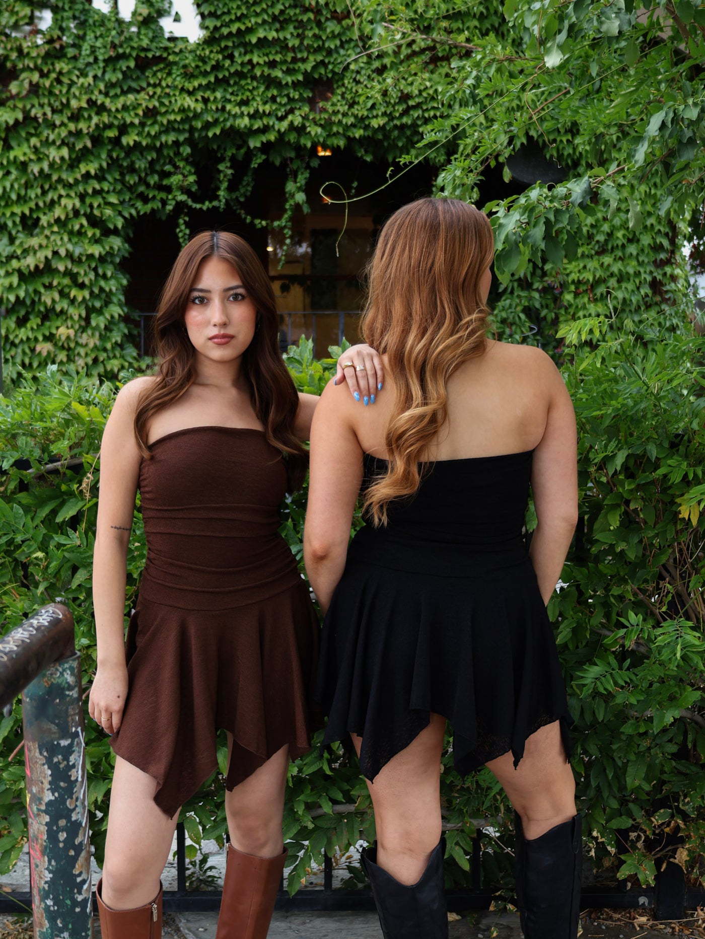Two women wearing sleeveless dresses standing in front of green foliage.