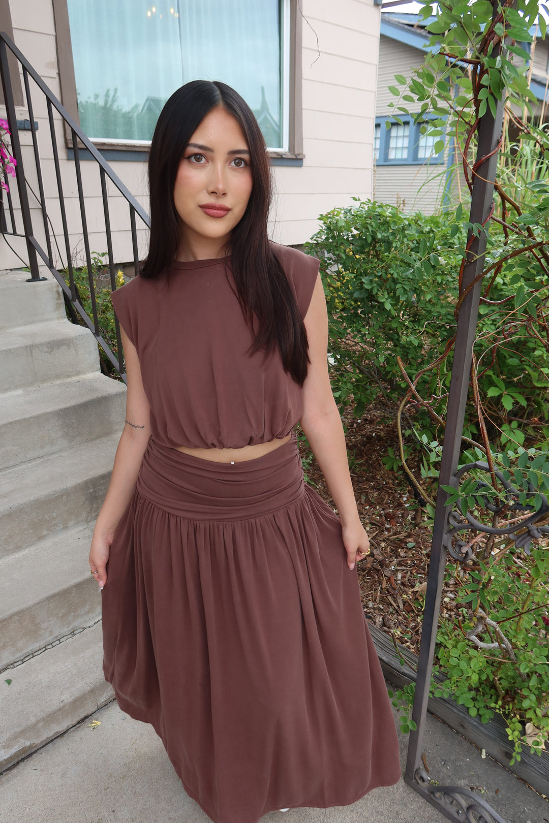 A woman wearing a brown two-piece dress with a sleeveless crop top and a pleated skirt, standing on outdoor concrete steps with green plants and a metal railing in the background.