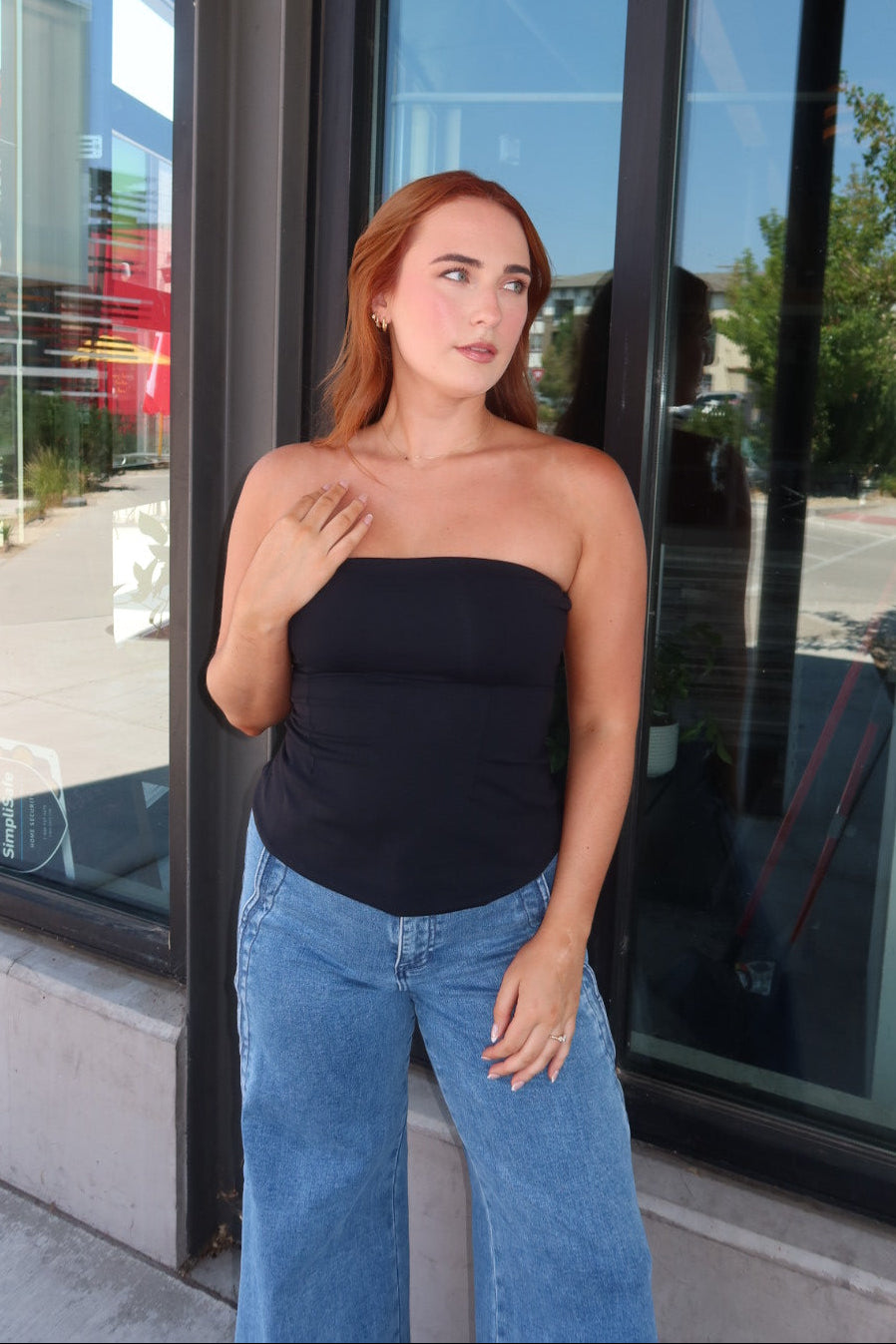 A woman with red hair stands in front of a storefront window wearing a black strapless tube top and light blue wide-leg jeans.
