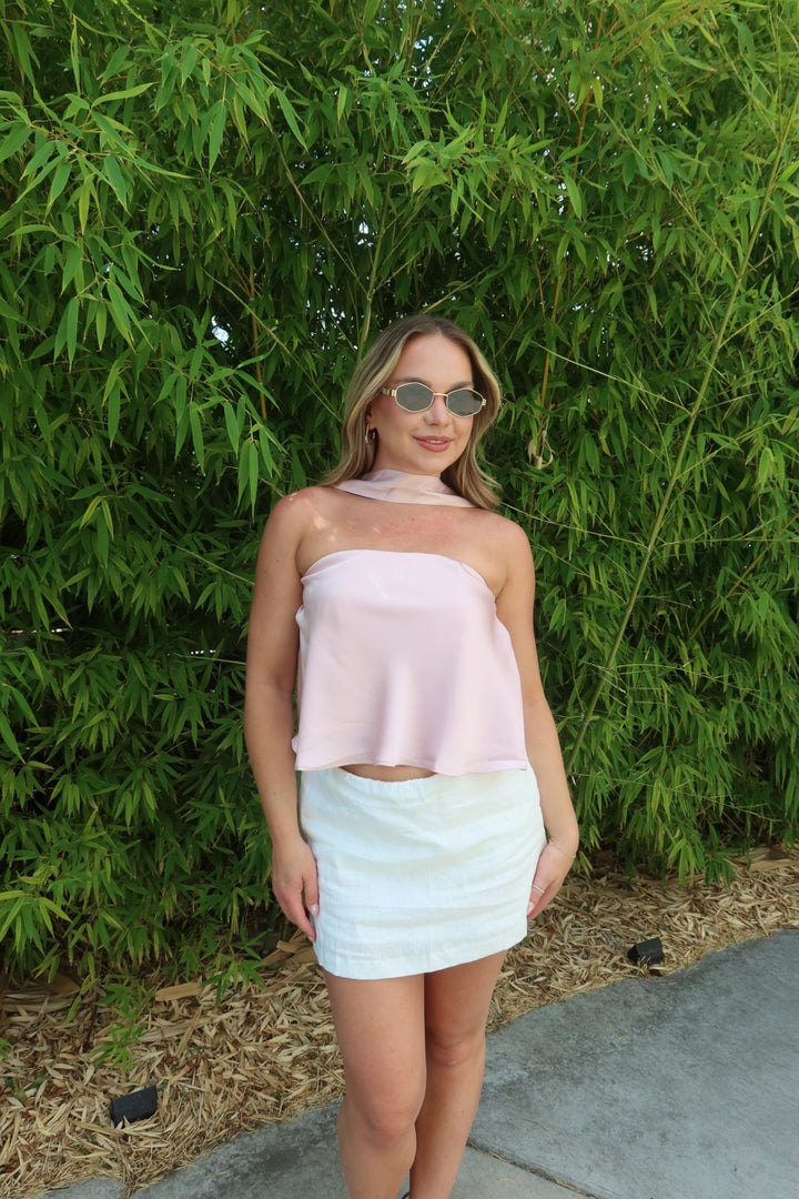 A woman wearing a light pink strapless tube top and a white striped mini skirt, standing outdoors in front of a dense green bamboo wall.