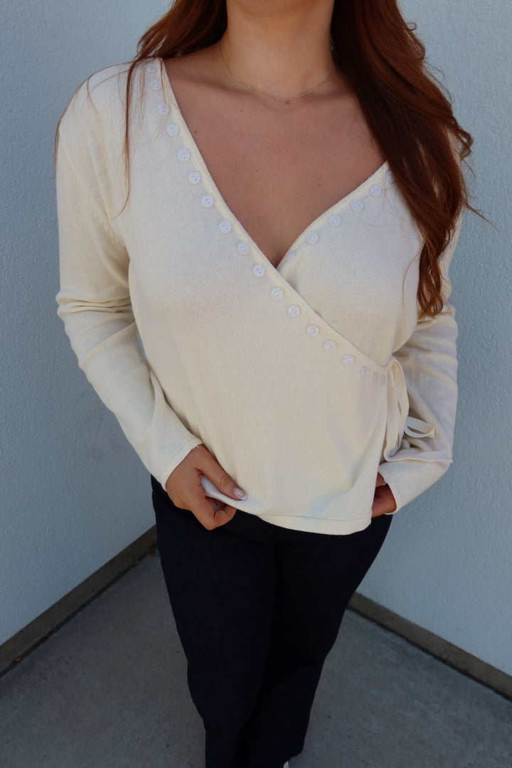 A woman wearing a cream-colored long-sleeve wrap-style top with white buttons down the front, paired with black pants, standing against a light gray wall.