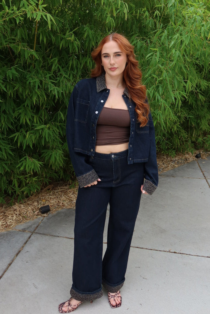 A woman with long red hair stands outdoors on a concrete path in front of a dense green bamboo wall, wearing a dark blue denim jacket with gray knit trim over a brown crop top and matching dark blue wide-leg jeans.
