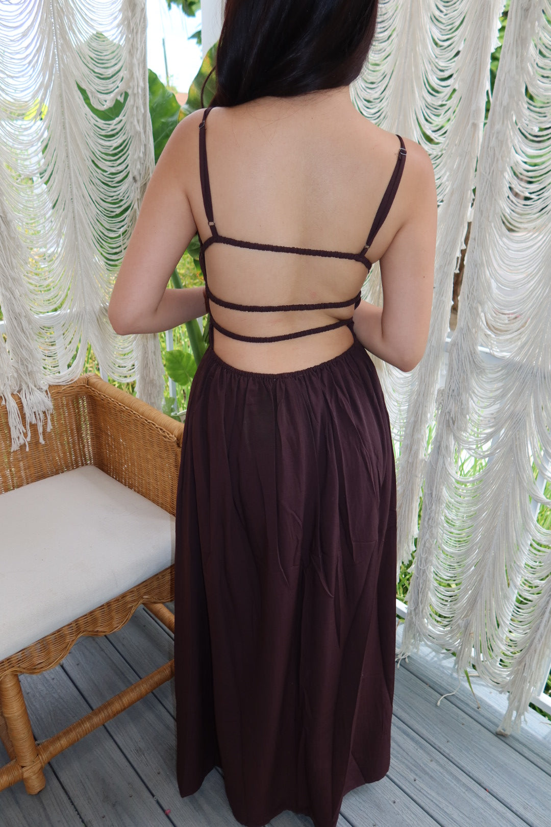 A woman viewed from behind wearing a dark brown maxi dress with thin spaghetti straps and multiple horizontal back straps, standing on a wooden deck in front of white macrame curtains.