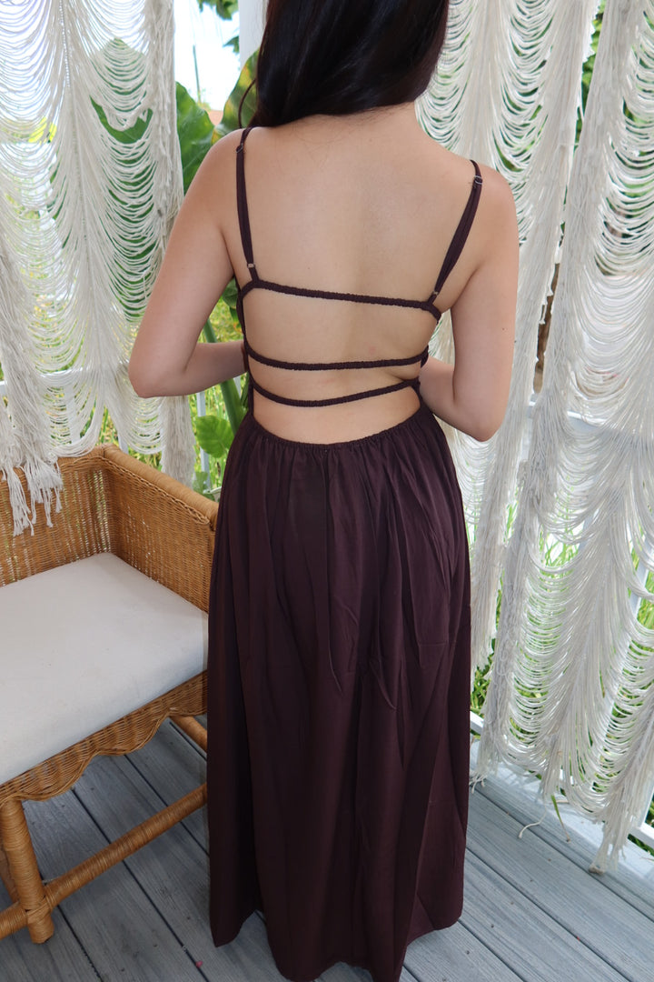 A woman viewed from behind wearing a dark brown maxi dress with thin spaghetti straps and multiple horizontal back straps, standing on a wooden deck in front of white macrame curtains.