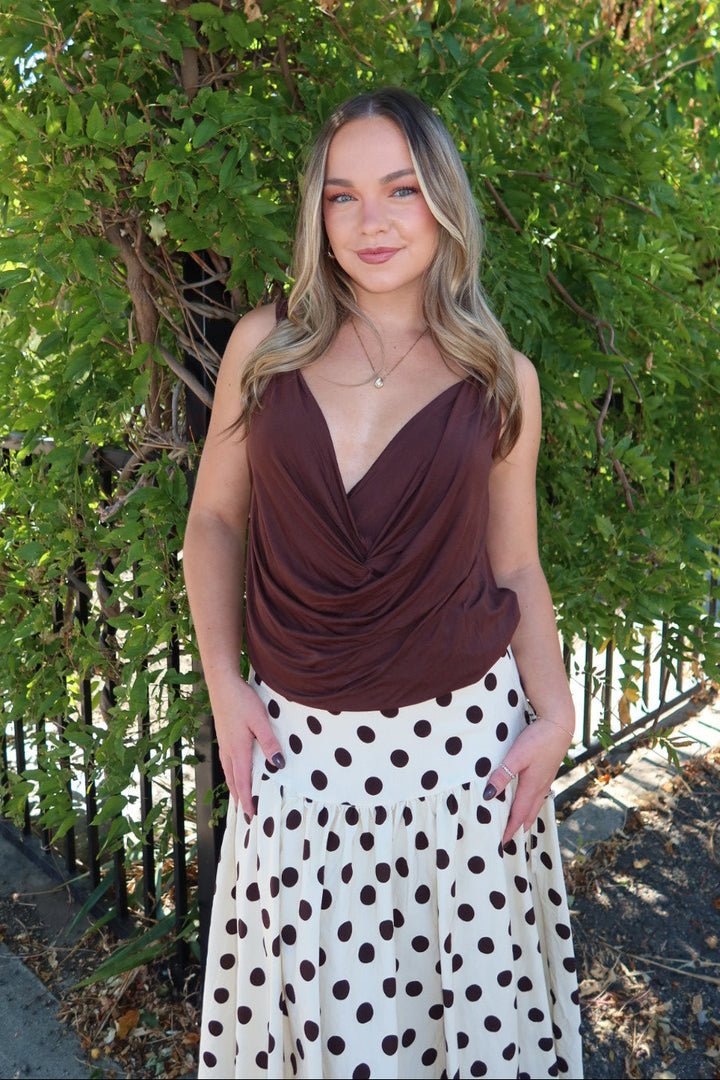 A woman standing outdoors in front of green foliage, wearing a dark brown draped sleeveless top and a white skirt with dark brown polka dots.