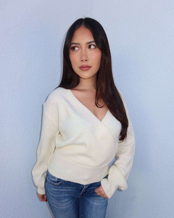 A woman with long dark hair wearing a cream-colored wrap-style knit sweater and blue jeans, standing against a light blue textured wall.