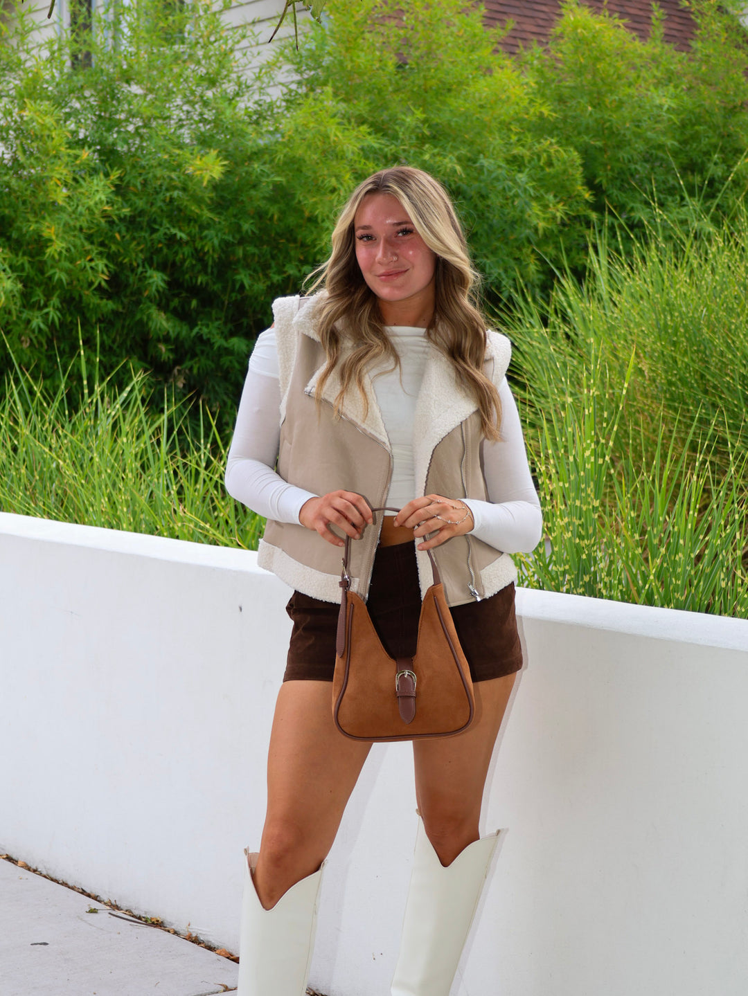 Woman wearing a beige vest, white shirt, brown shorts, and white boots with a brown bag outdoors.