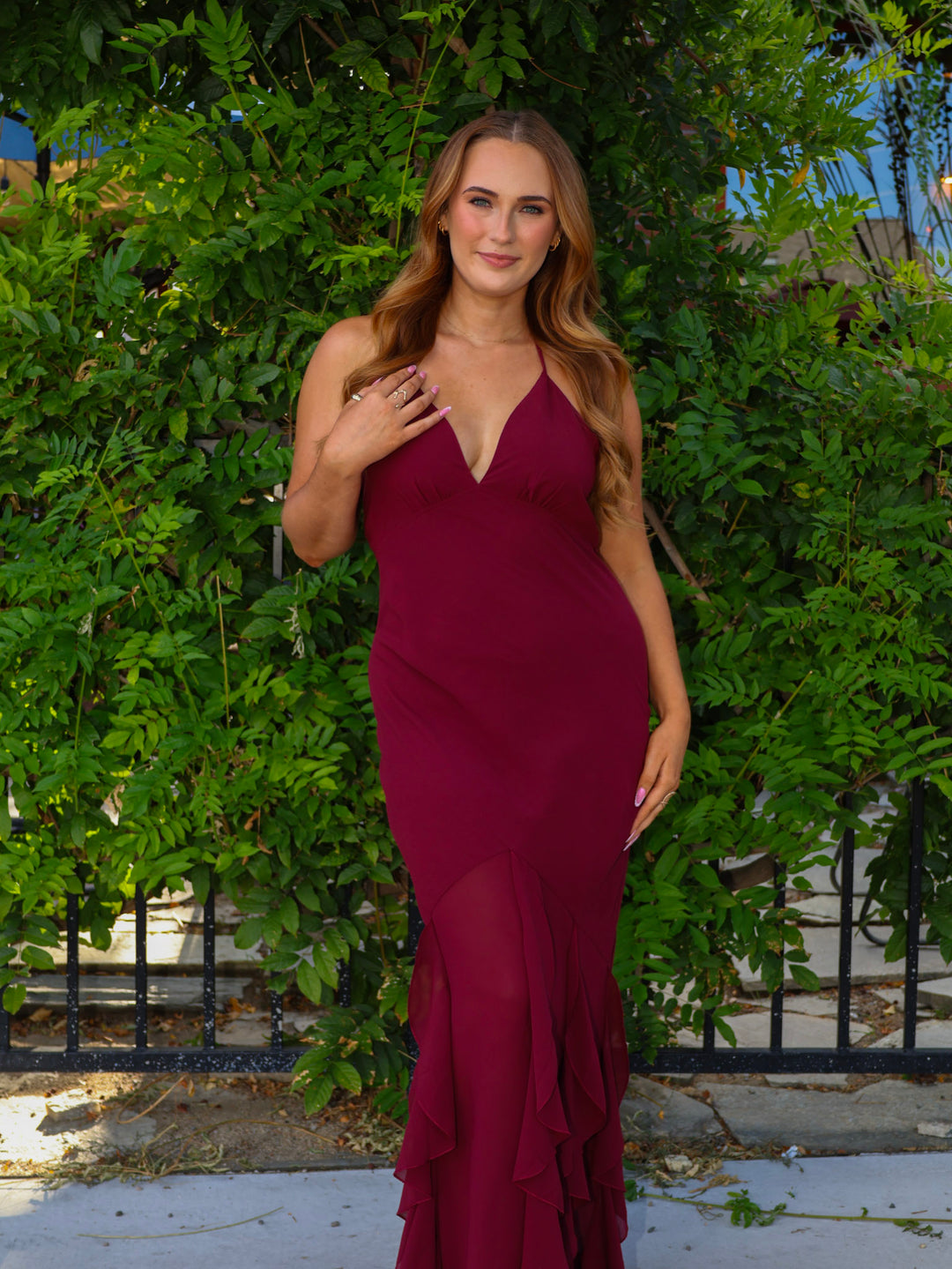 Woman in a burgundy evening gown standing against a green hedge.