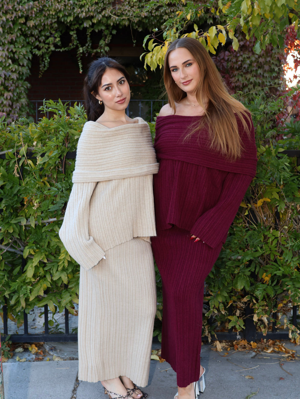 Two women in stylish outfits standing outdoors with greenery in the background