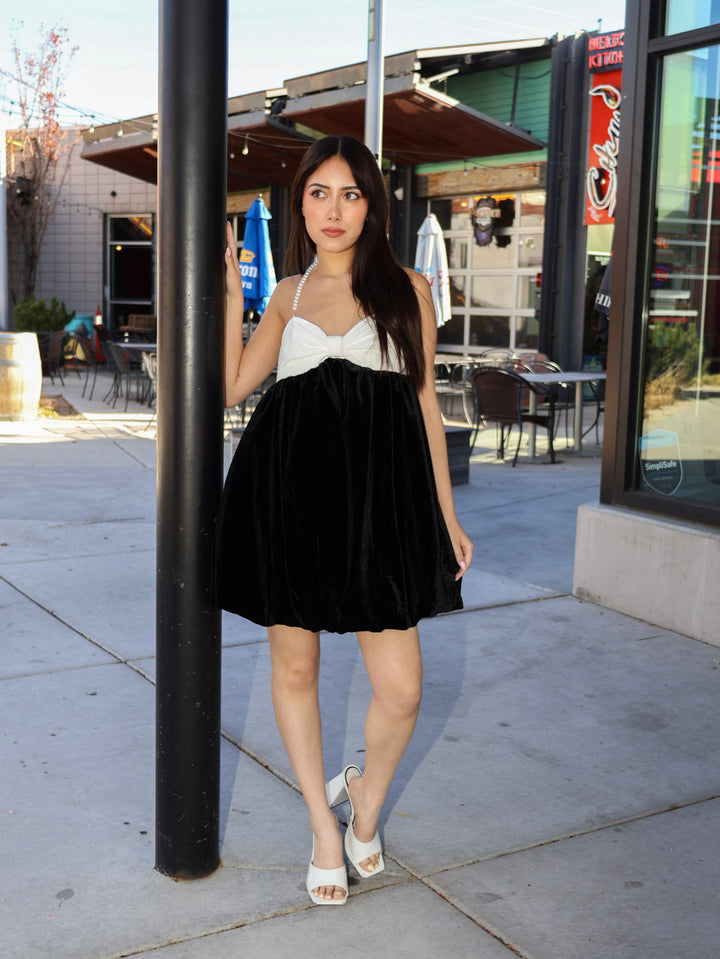 Woman in a black and white dress standing next to a pole on a sidewalk.