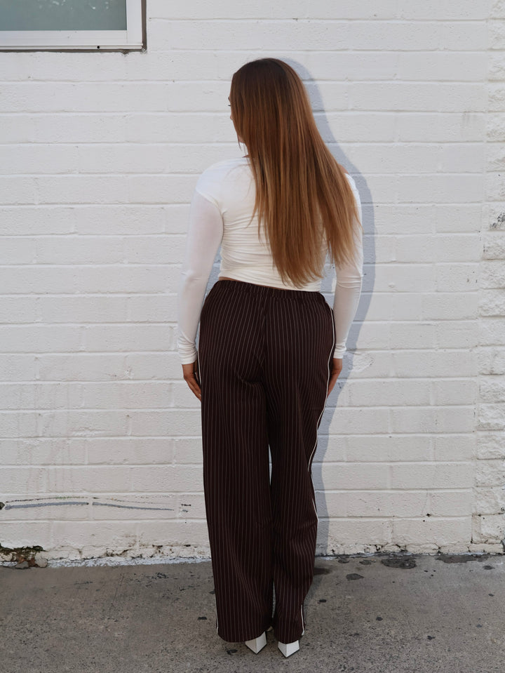 Person wearing a white top and brown pinstripe pants against a white brick wall.