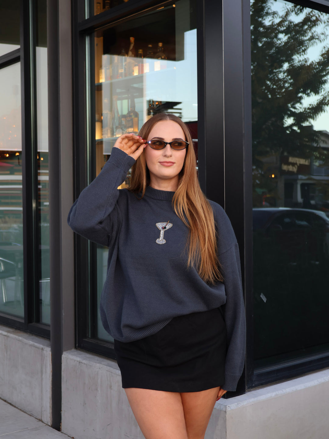 Woman wearing a dark gray sweatshirt with a logo and sunglasses, standing in front of a building.