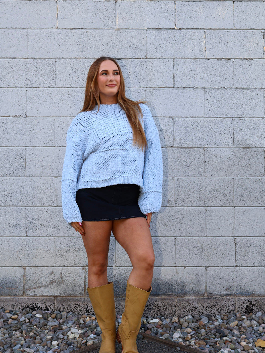 Woman wearing a light blue sweater and black skirt with yellow boots against a gray brick wall.