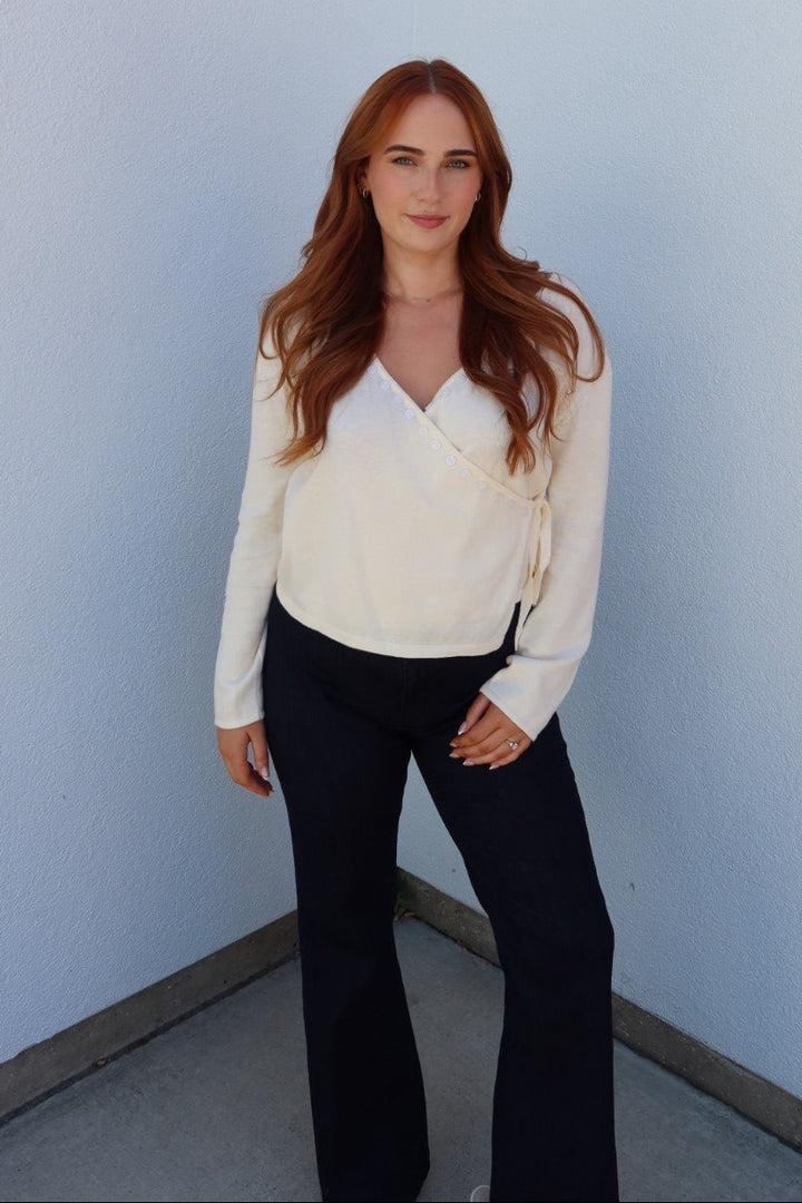 A woman with long red hair stands in a white and orange architectural corner, wearing a cream-colored wrap-style long-sleeve top and black pants.