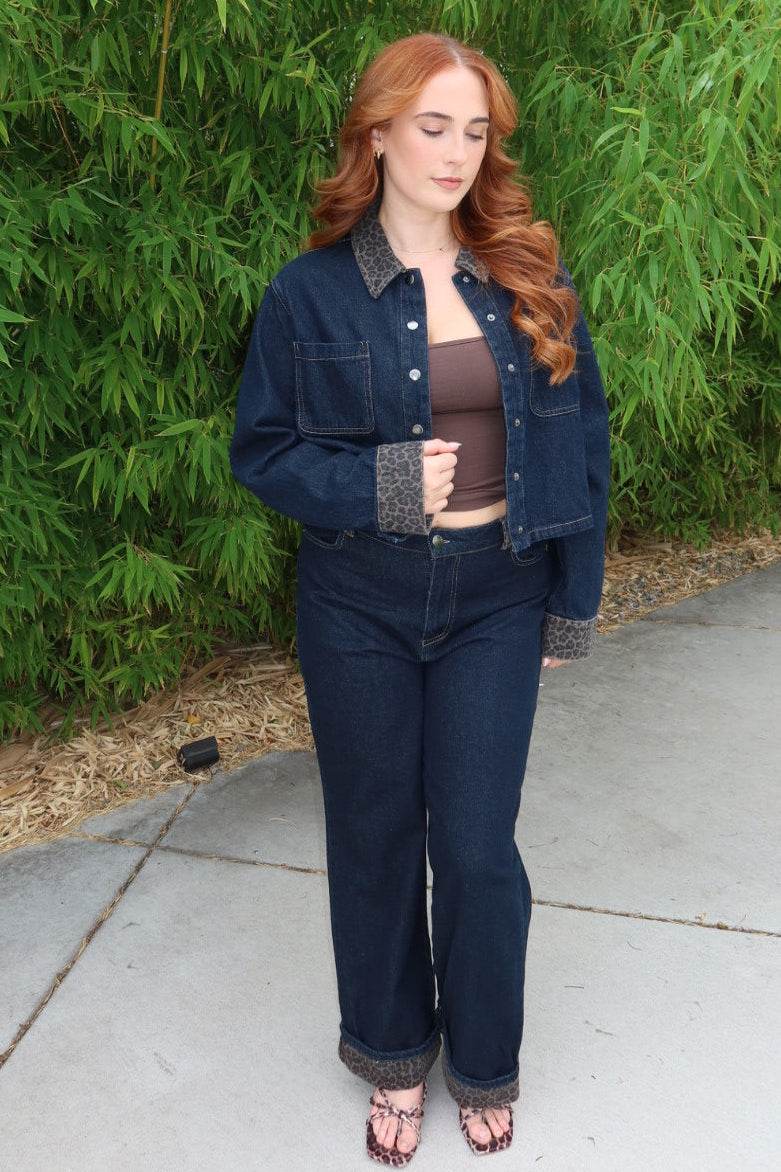 A woman with long red hair stands outdoors on a concrete walkway in front of a dense green bamboo hedge. She is wearing a dark blue denim jacket and matching dark blue jeans over a brown ribbed tank top, paired with dark sandals.