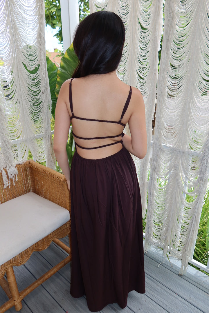 A woman viewed from behind wearing a dark plum maxi dress with thin strappy back details, standing on a porch in front of white macrame curtains.