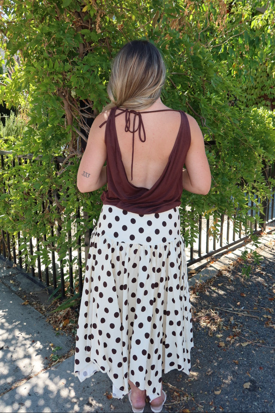 A woman viewed from behind wearing a brown halter top and a white midi skirt with dark brown polka dots, standing outdoors in front of a green hedge and black metal fence.
