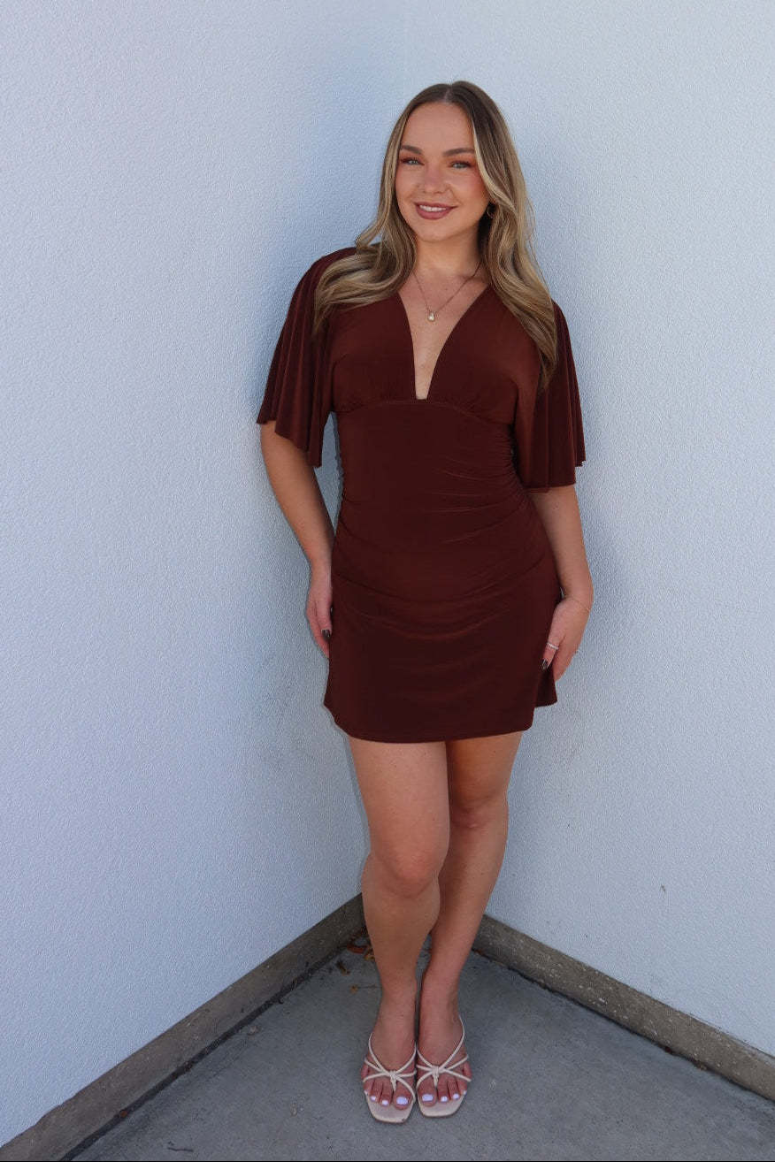 A woman modeling a short brown mini dress with short flutter sleeves and a deep V-neckline, standing against a white wall corner.