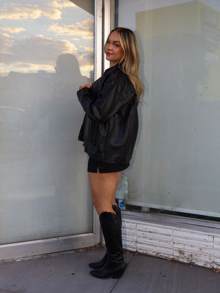 Person wearing a black outfit standing near a glass wall with a reflection.