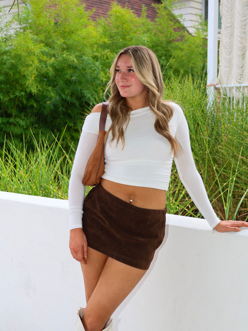 Woman in a white long-sleeve top and brown skirt standing outdoors with greenery in the background