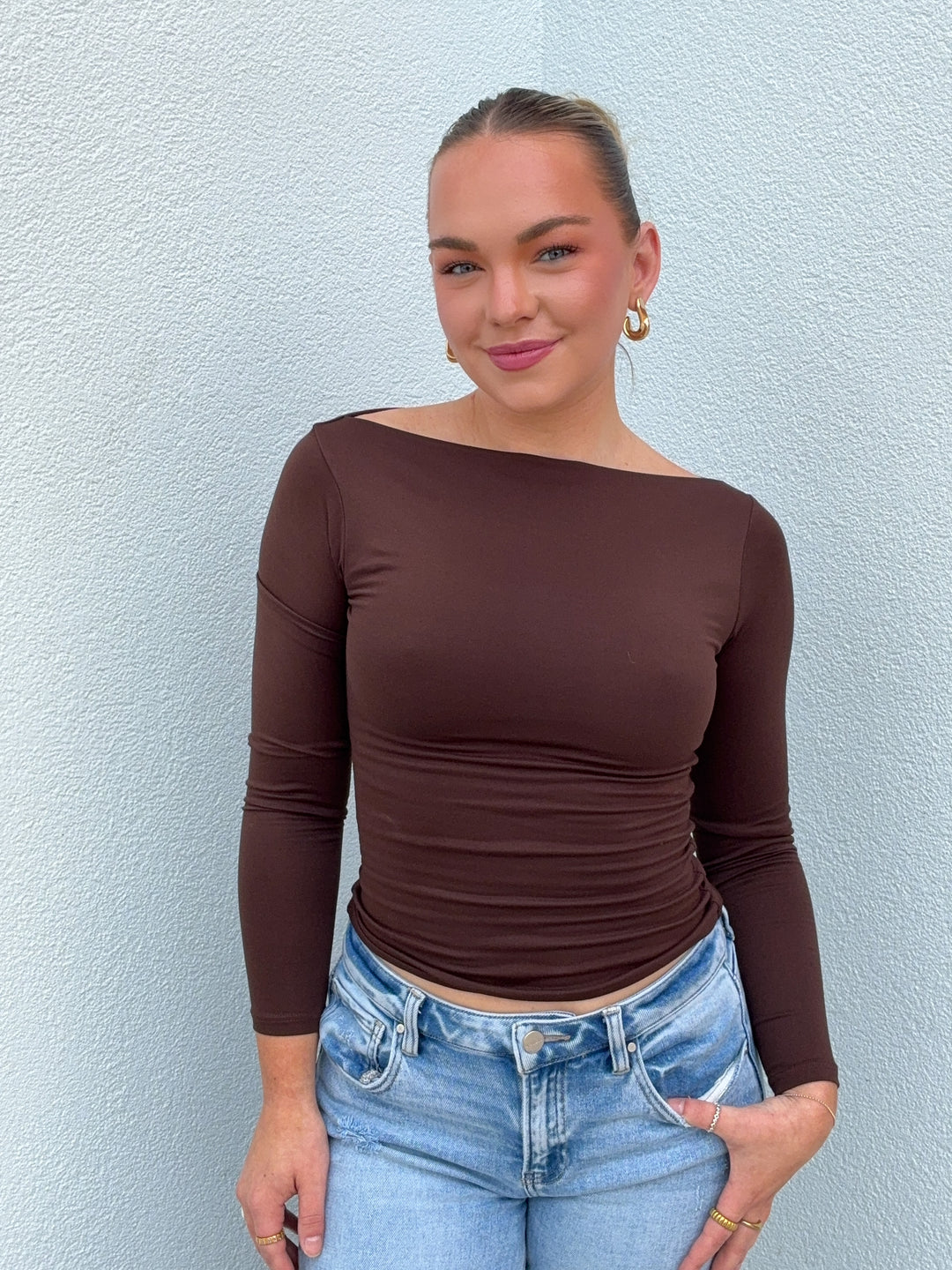 Woman wearing a brown long-sleeve top and blue jeans against a light blue wall.