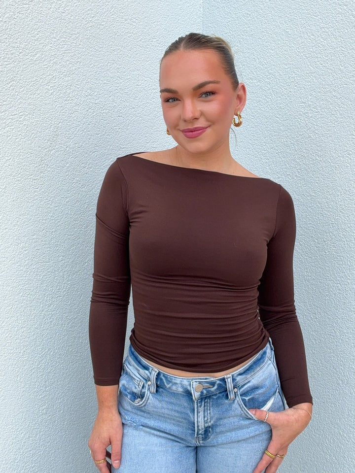 Woman wearing a brown long-sleeve top and blue jeans against a light blue wall.