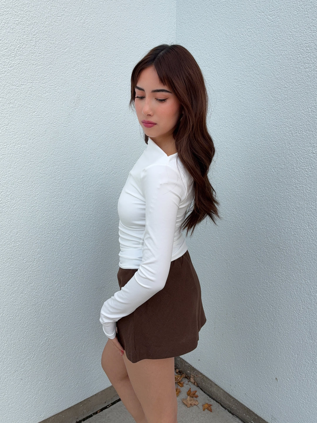 Woman wearing a white long-sleeve top and brown skirt against a light blue wall.
