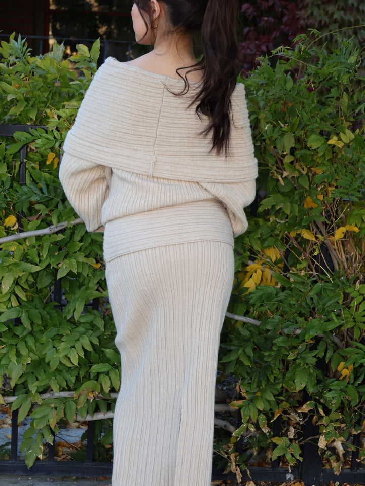 Person wearing a cream ribbed sweater and skirt standing in front of green foliage.